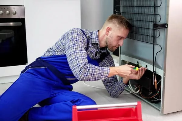 Técnico arrodillado reparando la parte trasera de un frigorífico junto a una caja de herramientas roja